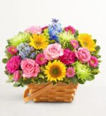 A vibrant and dense floral arrangement in a woven light-brown wooden basket, featuring pink roses, yellow sunflowers, purple carnations, lime green chrysanthemums, and blue delphiniums.