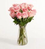 A dozen long-stemmed light pink roses arranged in a clear fluted glass vase against a plain white background.