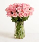 A premium bouquet of long-stemmed pink roses arranged in a tall, clear glass fluted vase against a plain white background.
