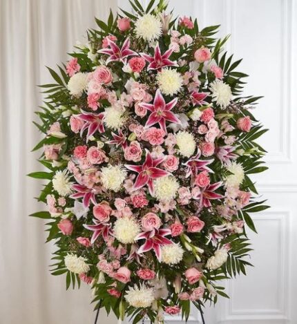 A large, lush funeral standing spray featuring a dense mix of pink Stargazer lilies, soft pink roses, white chrysanthemums, and pink carnations accented with rich green palm fronds and foliage.