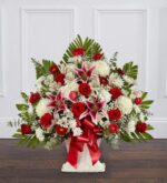 A large, fan-shaped floral arrangement in a white floor basket featuring red roses, pink and white lilies, white chrysanthemums, and green palm fronds, accented with a large red ribbon.