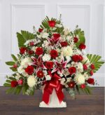 A large, fan-shaped floral arrangement in a white pedestal vase featuring deep red roses, pink and white stargazer lilies, white chrysanthemums, and lush green palm leaves, accented with a large red satin bow.