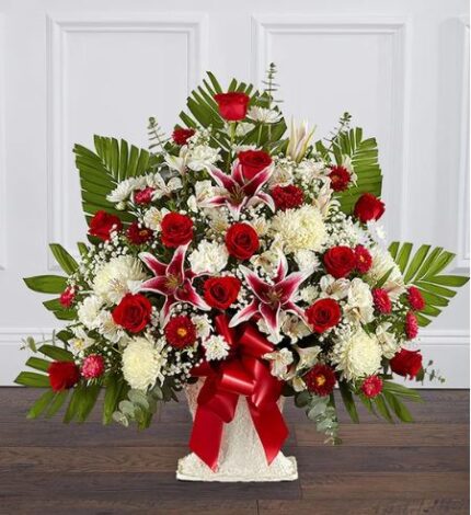 A large, fan-shaped floral arrangement in a white pedestal vase featuring deep red roses, pink and white stargazer lilies, white chrysanthemums, and lush green palm leaves, accented with a large red satin bow.