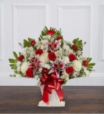 A large, fan-shaped floral arrangement in a white pedestal basket featuring red roses, pink and white stargazer lilies, white chrysanthemums, and lush greenery with a large red satin bow.