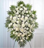 A large, elegant funeral standing spray featuring a dense arrangement of white flowers including roses, lilies, and carnations, accented with lush green palm fronds and filler greenery, displayed on a wire tripod easel against a neutral white wall.