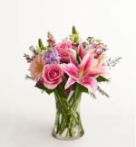 A beautiful floral arrangement in a clear glass vase featuring pink lilies, roses, and gerbera daisies with purple accents and green foliage.
