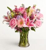 A lush and premium floral arrangement in a glass vase featuring a mix of pink roses, stargazer lilies, pink gerbera daisies, and purple stock flowers against a plain white background.