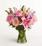 A lush floral arrangement in a clear glass vase featuring a mix of pink roses, a vibrant stargazer lily, peach gerbera daisies, and purple stock flowers against a plain white background.