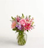 A lush and elegant floral arrangement in a clear glass vase featuring pink roses, pink oriental lilies, pink gerbera daisies, and purple stock flowers against a plain white background.