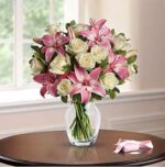 A bouquet of pink stargazer lilies and white roses arranged in a clear glass vase, placed on a round wooden table with a pink ribbon nearby and soft window light in the background.