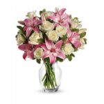 Elegant bouquet of pink stargazer lilies and white roses with green foliage in a clear glass vase, arranged against a white background.