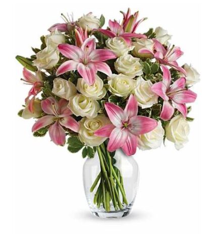 Elegant bouquet of pink stargazer lilies and cream-colored roses with green foliage in a clear glass vase