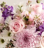 A lush and colorful floral bouquet featuring soft pink roses, light purple chrysanthemums, white daisies, and purple stocks.