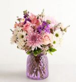 A lush floral arrangement featuring pink roses, purple stock flowers, and white daisies in a textured purple glass vase.