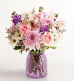 A lush mixed flower bouquet featuring pink roses, purple stocks, and light pink chrysanthemums in a textured lavender glass vase.