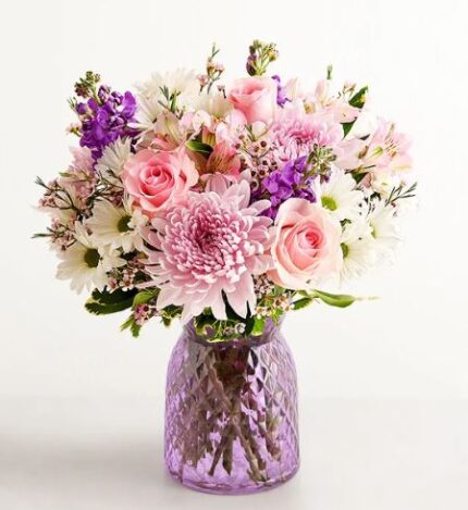 A lush mixed flower bouquet featuring pink roses, purple stocks, and light pink chrysanthemums in a textured lavender glass vase.