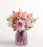 A vibrant floral arrangement featuring pink roses, chrysanthemums, and alstroemeria in a textured purple glass vase against a plain white background.
