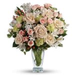 Elegant floral arrangement in a clear glass vase with blush and cream roses, pink spray roses, white alstroemeria, light green hydrangea, and pink hypericum berries, accented with dark green foliage.