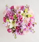 A lush top-down view of a round floral bouquet featuring a mix of pink roses, white lilies, light purple stocks, and pink chrysanthemums against a neutral light grey background.
