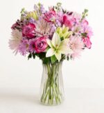 A lush and vibrant mixed flower bouquet in a clear glass vase, featuring purple roses, white lilies, and light pink chrysanthemums against a plain white background.