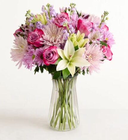 A lush and vibrant mixed flower bouquet in a clear glass vase, featuring purple roses, white lilies, and light pink chrysanthemums against a plain white background.