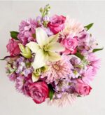 A vibrant top-down view of a floral bouquet featuring a prominent white lily surrounded by pink roses, purple stocks, and lavender chrysanthemums.