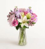 A lush floral arrangement in a clear fluted glass vase featuring white lilies, pink and lavender roses, purple stock flowers, and light pink chrysanthemums against a plain white background.