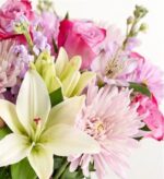 A vibrant close-up of a floral bouquet featuring a large white lily, pink roses, light pink chrysanthemums, and purple alstroemeria against a white background.