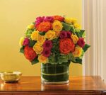 A round, colorful floral arrangement in a clear glass cylinder vase on a wooden table. The bouquet features vibrant orange, yellow, and hot pink roses mixed with lime green hydrangeas, set against a soft yellow wall. A small gold bowl sits to the left.