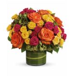 Round floral bouquet in a short green glass vase featuring vibrant orange, yellow, and hot pink roses mixed with green hydrangea, viewed from the front on a white background.