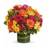 Vibrant mixed bouquet of orange, hot pink, and yellow roses with green hydrangea accents in a round glass vase wrapped with green leaf lining