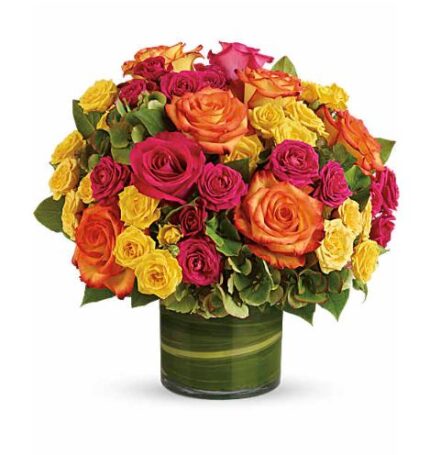 Vibrant mixed bouquet of orange, hot pink, and yellow roses with green hydrangea accents in a round glass vase wrapped with green leaf lining