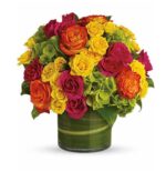 Round floral arrangement in a clear glass vase wrapped with green leaves. Features bright yellow, orange, and hot pink roses mixed with lime green hydrangeas.