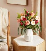 A vibrant floral arrangement in shades of blush pink, peach, and deep red, displayed in a textured white ceramic vase on a hexagonal marble-top wooden side table.
