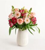 A lush floral arrangement featuring soft pink roses, peach gerbera daisies, and deep magenta alstroemeria in a white textured ceramic vase.
