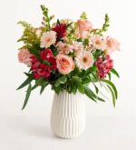 A lush floral bouquet featuring pink roses, light pink gerbera daisies, and deep pink alstroemeria, arranged in a white textured ceramic vase against a plain white background.