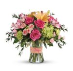 Elegant glass vase filled with hot pink roses, peach lilies, pink alstroemeria, blush spray roses, and green hydrangea, accented with greenery and tied with a peach ribbon.