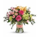 A glass vase with a peach ribbon holds a bouquet of pink roses, peach lilies, light pink alstroemeria, pale pink spray roses, green hydrangeas, and filler greenery.
