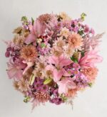 A top-down view of a lush, round floral bouquet featuring a variety of pink and peach flowers, including lilies, chrysanthemums, and snapdragons, arranged densely against a light gray background.