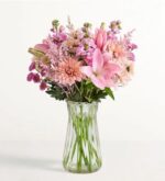 A lush floral arrangement in a clear fluted glass vase, featuring a mix of pink lilies, peach chrysanthemums, purple stock flowers, and light pink astilbe against a plain white background.