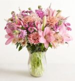 A lush and elegant bouquet of pink lilies, peach chrysanthemums, and purple stock flowers in a clear, fluted glass vase against a white background.