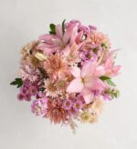 A top-down view of a lush "Blushing Glow" floral bouquet featuring a mix of pink lilies, peach chrysanthemums, and purple spray asters against a neutral off-white background.