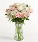 A pastel flower bouquet in a clear glass vase. Features light pink roses, white alstroemeria, pale pink carnations, and white chrysanthemums with green foliage, set against a white background.
