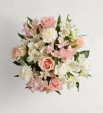 Top-down view of a round floral bouquet with blush pink roses, white carnations, and white and light pink alstroemeria lilies arranged against a light neutral background.