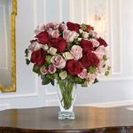 A large, round bouquet of deep red and soft pink roses with small white accent flowers and green foliage, arranged in a clear glass vase on a polished wooden table. The background shows a light gray paneled wall with gold-framed mirror and wall sconce.