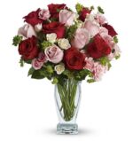 A romantic mixed rose bouquet in a clear glass vase, featuring deep red roses, soft pink roses, and small white spray roses, accented with lush green foliage.