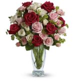 A lush bouquet of red, pink, and white roses with small green filler flowers arranged in a clear glass vase with long green stems visible.
