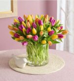 A vibrant and dense bouquet of assorted tulips in shades of purple, yellow, and orange, arranged in a clear cylindrical glass vase on a pink table with a decorative bird figurine.
