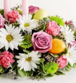 A vibrant Easter floral centerpiece featuring white daisies, purple roses, pink carnations, and green chrysanthemums, decorated with yellow and orange Easter eggs and pink candles.
