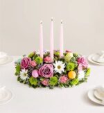 A lush, oval-shaped Easter floral centerpiece featuring pink roses, white daisies, green chrysanthemums, and pink carnations, accented with three tall pink taper candles and colorful Easter eggs on a white tablecloth.